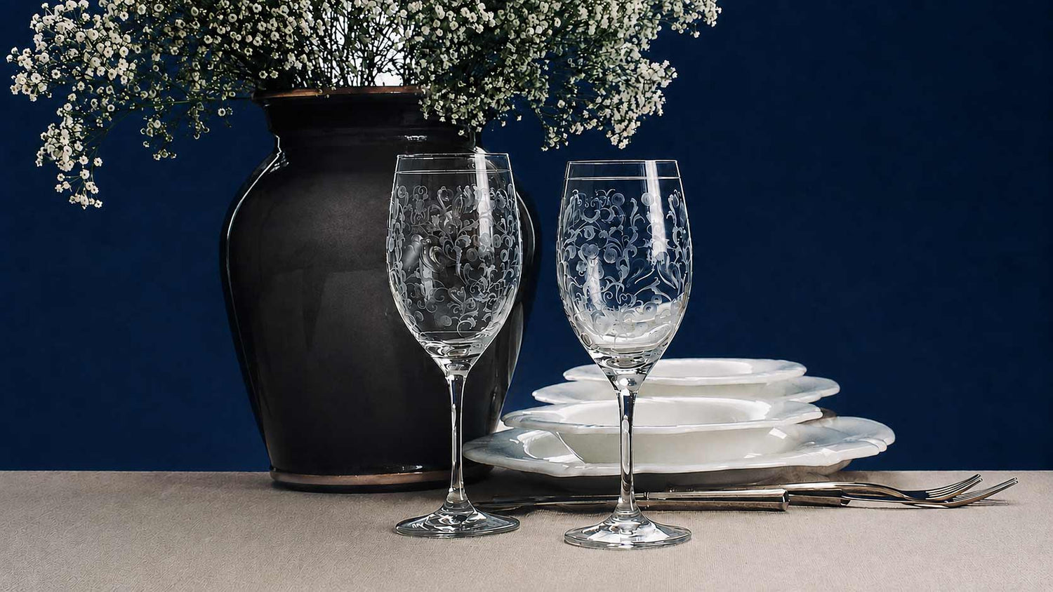 Zwei Weingläser aus Kristall mit reicher Handgravur vor einer dunklen Vase mit Blumen